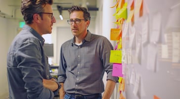 Two engineers having a casual discussion in front of a wall covered with colorful sticky notes during a problem-solving session.